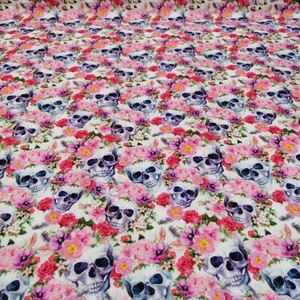 Puede incluir: Tela blanca con un patrón repetido de flores coloridas y calaveras. Las calaveras son azules y moradas con ojos blancos y están rodeadas de flores rosas, moradas y blancas.