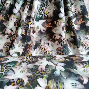 Op de afbeelding: Stof met een bloemen- en dierenprint. Het toont witte lelies met roze accenten tegen een donkere achtergrond met een luipaardprint in tinten beige, bruin en geel. De stof is opgevouwen.