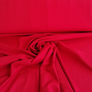 Op de afbeelding: Een close-up van een felrode stof, gevouwen en gedrapeerd in een zachte, vloeiende manier. De stof heeft een gladde, gelijkmatige textuur en een rijke, levendige kleur.