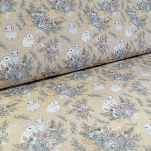 Puede incluir: Una tela beige con un patrón repetido de conejos blancos sentados en ramos de flores. Los ramos están hechos de flores azules, rosas y blancas.