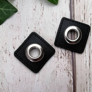 Può includere: Due quadrati in pelle nera con occhielli in metallo argentato. Gli occhielli sono rotondi e hanno una finitura liscia e lucida.