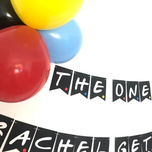 May include: Black and white banner with the text "The One Where Rachel Gets Married" in a white handwritten font. The banner is decorated with colorful dots. The banner is hanging from balloons in red, yellow, and blue.