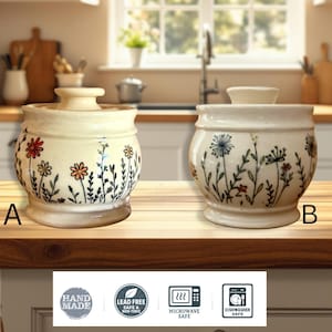 May include: Two off-white ceramic jars with lids, labeled A and B, featuring hand-painted wildflower designs in red, orange, and green. Below the jars, four icons indicate the items are handmade, lead-free, microwave-safe, and dishwasher-safe.