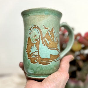 Puede incluir: Una taza de cerámica con un esmalte verde claro y una ilustración marrón de una escena de lago con un dinosaurio, árboles y montañas. La taza tiene un asa curva y un borde ancho.