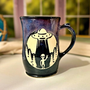 May include: A black and purple ceramic mug with a white design of an alien waving and a UFO flying over a forest.
