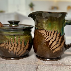 May include: A brown and green ceramic sugar bowl with a lid and a matching creamer with a fern design. The sugar bowl has a wooden spoon.