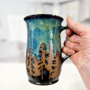 Puede incluir: Una taza de cerámica azul y verde con una silueta marrón de una escena de bosque. La taza tiene un asa y un fondo redondeado.