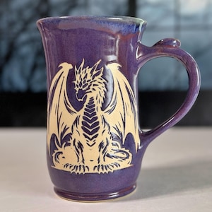 May include: A tall, purple ceramic mug featuring a cream-colored, etched graphic of a dragon with spread wings on the front. The mug has a smooth, glossy finish, a flared rim, and a curved handle with a small thumb rest at the top.