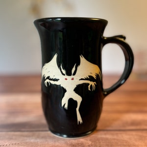 May include: Black ceramic mug with a white cryptid design featuring red eyes. The mug has a curved handle and a flared top. The design is a stylized illustration of a moth-like creature. The mug is on a wooden surface.