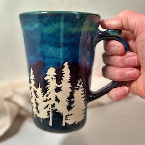 May include: A tall ceramic mug with a forest silhouette design. The mug features a gradient of blue and green hues, with a white tree line. The handle is dark blue. The mug is held by a hand.