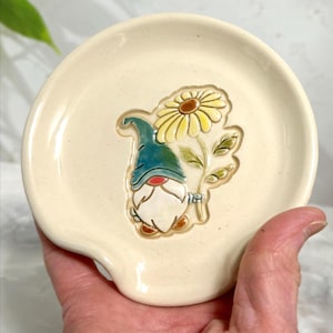 Puede incluir: Apoyacucharas de cer&aacute;mica color crema con un dise&ntilde;o caprichoso de gnomo y margarita. El gnomo lleva un gorro verde azulado y sostiene una margarita amarilla. El apoyacucharas tiene forma curva con un peque&ntilde;o labio.