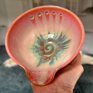 May include: A pink ceramic bowl with a swirl design in shades of blue, green, and brown. The bowl has a pour spout and five holes around the rim.