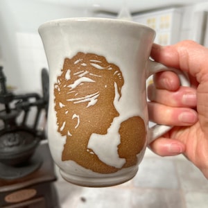 May include: White ceramic mug with a brown silhouette of a woman and child. The mug has a handle and a rounded shape.