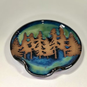 May include: A ceramic spoon rest with a forest silhouette design. The spoon rest features a brown tree line against a backdrop of green and blue glaze. The rim is dark blue.