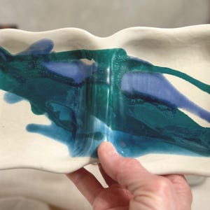 Puede incluir: Una fuente de cerámica blanca con borde ondulado, dividida en dos secciones. La fuente está decorada con trazos abstractos de pintura azul y verde azulado. La fuente está sostenida por una persona.