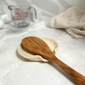 Puede incluir: Una cuchara de madera descansa sobre un reposacucharas de cer&aacute;mica color crema. La cuchara es de color marr&oacute;n claro con vetas de madera visibles. Un vaso medidor est&aacute; en el fondo. Utensilios de cocina.