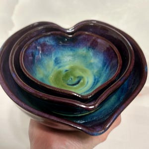 Puede incluir: Un juego de tres cuencos de cerámica en forma de corazón, anidados. Los cuencos presentan una vibrante paleta de colores púrpura intenso, verde azulado y verde, con un acabado brillante. Los cuencos están diseñados con una forma suave y curvada.
