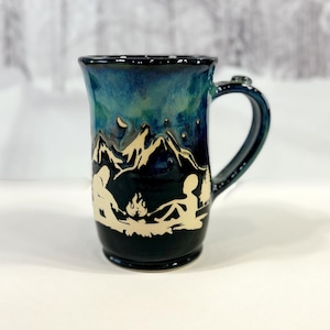 May include: A ceramic mug with a dark blue and teal glaze. The mug features a mountain scene with a campfire and figures carved into the surface. The handle is also dark blue. The mug is approximately 13 cm tall.
