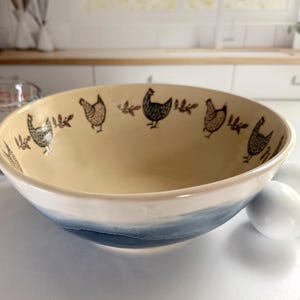 May include: A ceramic mixing bowl with a cream-colored interior featuring a repeating pattern of dark brown chickens and small leaves. The exterior of the bowl has a smooth, glossy finish with a deep blue gradient fading into a white base.