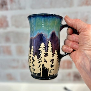 May include: A ceramic mug with a dark blue, purple, and green glaze. The mug has a silhouette of a forest with trees in a light beige colour.