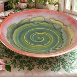 May include: A large, handmade ceramic bowl with a spiral design. The bowl features a color gradient from a reddish-orange rim to a green and blue center. The bowl is approximately 14 inches in diameter, perfect for serving.