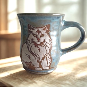 May include: A ceramic mug with a blue and grey mottled glaze featuring a detailed, etched illustration of a long-haired cat in white and brown tones. The mug has a sturdy handle and a slightly flared rim, showcasing a handcrafted appearance.