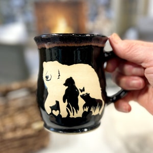 May include: Black ceramic mug with a bear family design. The mug features a large cream-colored bear with a heart, and two smaller black bears with a butterfly. The rim has a brown glaze.