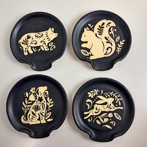 May include: Set of four black ceramic spoon rests, each featuring a different animal design in a cream color. The designs include a pig, squirrel, dog, and rabbit, with floral and leaf accents. These spoon rests are a kitchenware item.