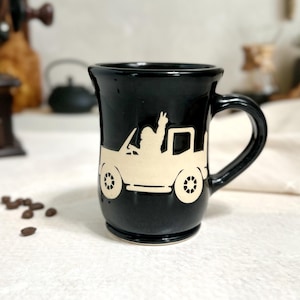 May include: Black ceramic mug with a cream-colored graphic of a vintage convertible with a person giving a peace sign. The mug has a curved handle and a slightly flared rim. Coffee beans are scattered near the mug.