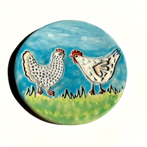 May include: A round ceramic tile featuring two chickens standing on a green grassy field against a bright blue sky. The left chicken has a white body with raised dots, while the right chicken is white with a red comb and textured wing details.