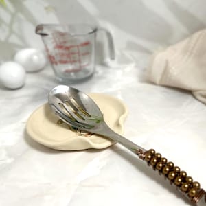 Puede incluir: Una cuchara ranurada de acero inoxidable descansa sobre un reposacucharas de cer&aacute;mica color crema con un dise&ntilde;o floral. El mango de la cuchara est&aacute; decorado con cuentas y alambre marrones. Un vaso medidor y esferas blancas est&aacute;n en el fondo.