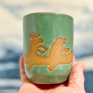May include: A green ceramic tumbler with a brown moose and bear design. The cylindrical cup has a glossy finish and is held in a hand. The moose and bear are side by side.
