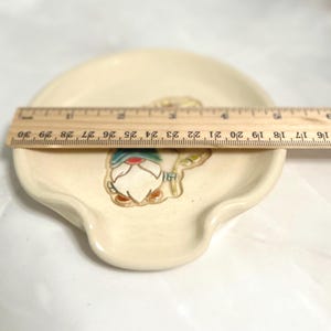 Puede incluir: Descansacucharas de cer&aacute;mica color crema con dise&ntilde;o de gnomo. El descansacucharas mide aproximadamente 25 cm de largo. El gnomo tiene un gorro azul, detalles rojos y barba blanca. El descansacucharas tiene forma curva con un peque&ntilde;o labio.