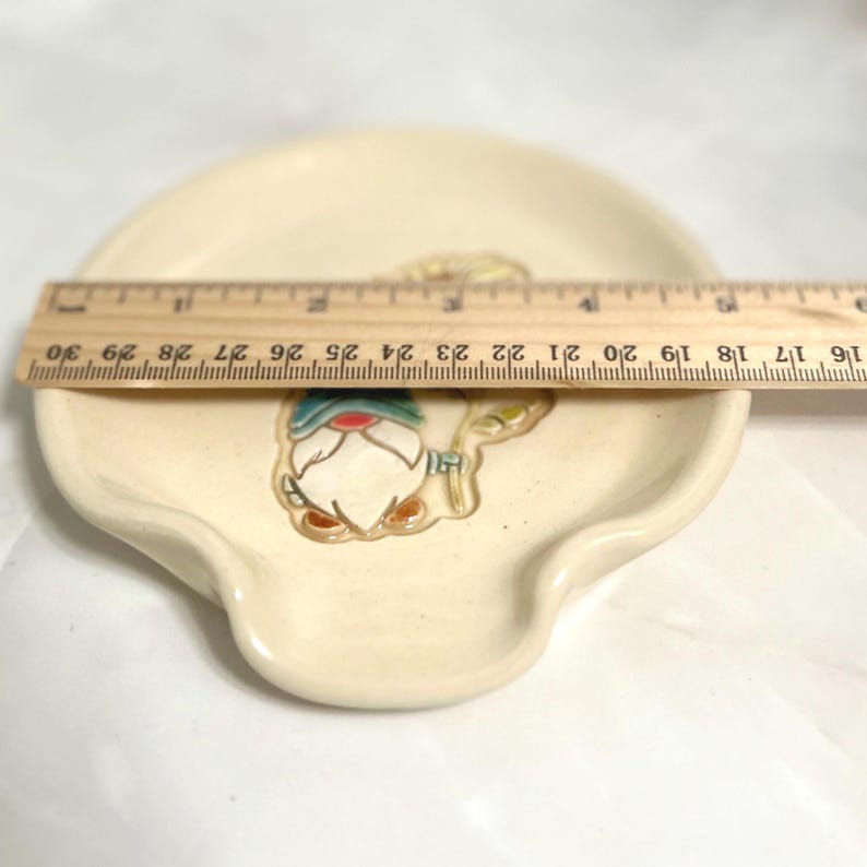 Puede incluir: Descansacucharas de cer&aacute;mica color crema con dise&ntilde;o de gnomo. El descansacucharas mide aproximadamente 25 cm de largo. El gnomo tiene un gorro azul, detalles rojos y barba blanca. El descansacucharas tiene forma curva con un peque&ntilde;o labio.