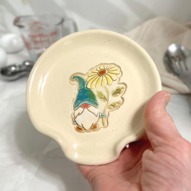Puede incluir: Apoyacucharas de cer&aacute;mica color crema con un dise&ntilde;o en relieve de un gnomo sosteniendo una margarita. El gnomo lleva un gorro azul y tiene barba blanca. La margarita es amarilla con centro marr&oacute;n. El apoyacucharas tiene forma curva.