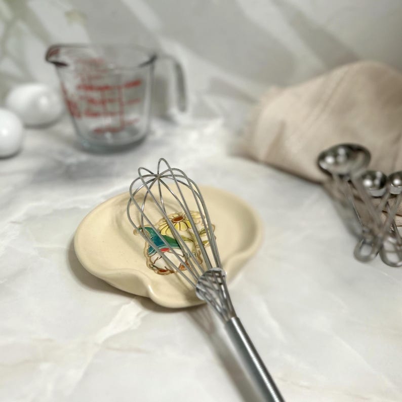 Puede incluir: Un batidor de acero inoxidable descansa sobre un reposacucharas de cer&aacute;mica color crema con un dise&ntilde;o floral. Una jarra medidora de vidrio y cucharas medidoras est&aacute;n en el fondo. Utensilios y accesorios de cocina.