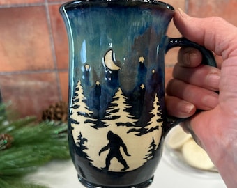 Può includere: Una tazza in ceramica con un design di Bigfoot. La tazza presenta una glassa blu scuro e verde acqua con una silhouette di Bigfoot che cammina davanti a una foresta e una luna crescente. La tazza ha un manico grande.