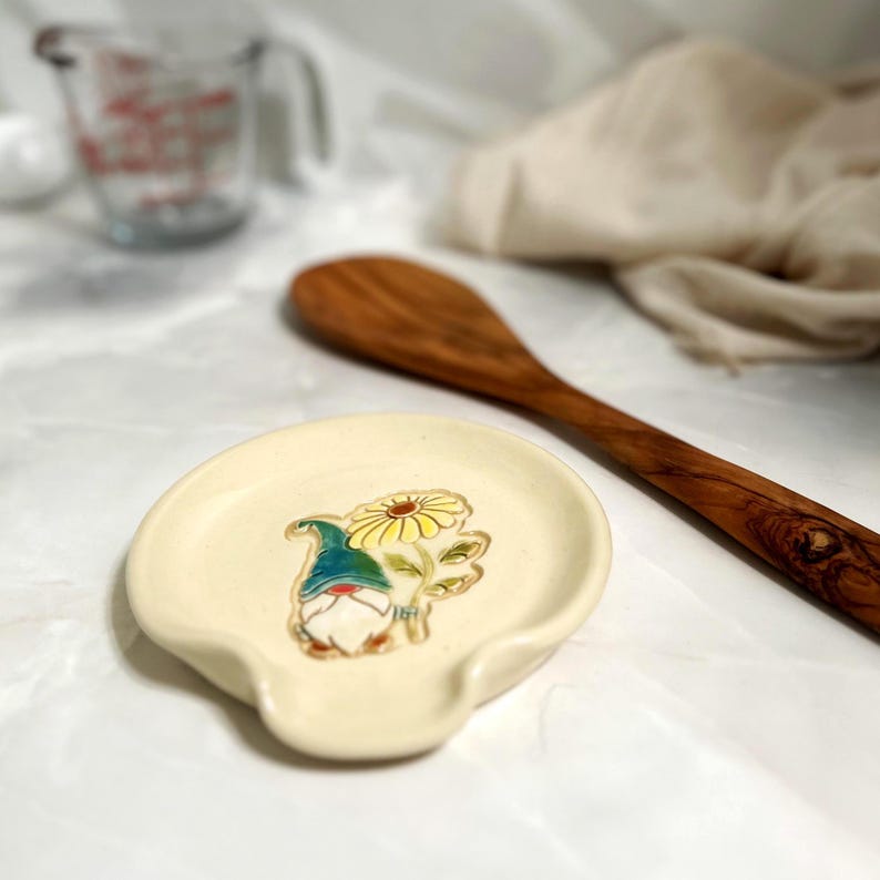 Puede incluir: Un reposacucharas de cer&aacute;mica color crema con un dise&ntilde;o caprichoso de gnomo y flor. El gnomo lleva un sombrero azul y sostiene una flor amarilla. Una cuchara de madera de olivo y una taza medidora de vidrio est&aacute;n en el fondo.