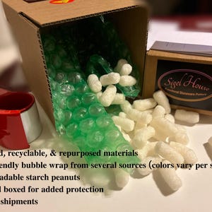 Puede incluir: Una caja de cart&oacute;n con pl&aacute;stico de burbujas verde y cacahuetes de embalaje blancos que se derraman. Se ve una caja m&aacute;s peque&ntilde;a con el logotipo de "Sigel House". Un dispensador de cinta adhesiva roja est&aacute; a la izquierda. La imagen destaca el embalaje ecol&oacute;gico.