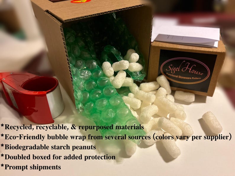 Puede incluir: Una caja de cart&oacute;n con pl&aacute;stico de burbujas verde y cacahuetes de embalaje blancos que se derraman. Se ve una caja m&aacute;s peque&ntilde;a con el logotipo de "Sigel House". Un dispensador de cinta adhesiva roja est&aacute; a la izquierda. La imagen destaca el embalaje ecol&oacute;gico.