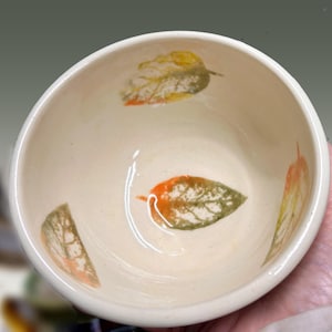 May include: Cream ceramic bowl featuring leaf designs in green, orange, and yellow. The bowl has a rounded shape and a smooth, glossy finish. The leaf patterns are evenly spaced around the interior of the bowl.