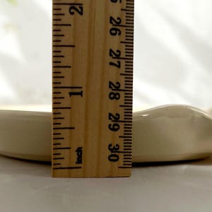 Puede incluir: Un peque&ntilde;o plato de cer&aacute;mica blanquecina con un borde ondulado. Una regla de madera se coloca junto al plato, mostrando una longitud de aproximadamente 7,6 cm. El plato tiene un dise&ntilde;o sencillo y elegante.