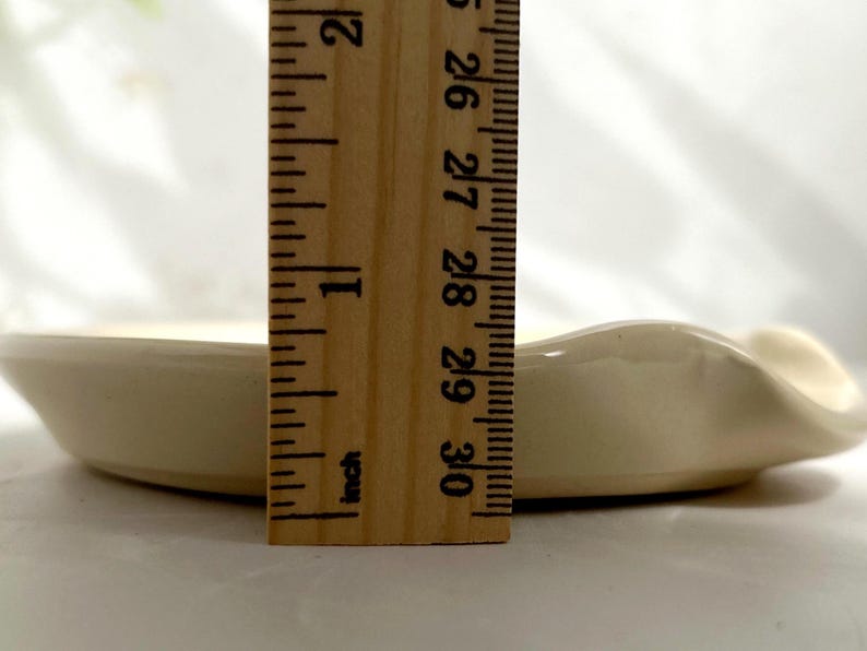 Puede incluir: Un peque&ntilde;o plato de cer&aacute;mica blanquecina con un borde ondulado. Una regla de madera se coloca junto al plato, mostrando una longitud de aproximadamente 7,6 cm. El plato tiene un dise&ntilde;o sencillo y elegante.