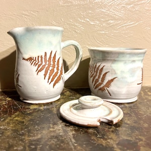 May include: A white ceramic creamer and sugar bowl set with a fern design. The creamer has a handle and the sugar bowl has a lid with a small pour spout.