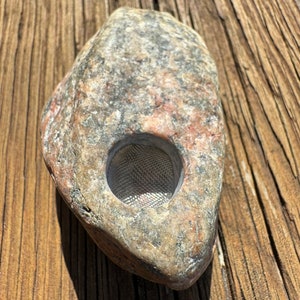 May include: A grey stone pipe with a metal screen in the bowl. The pipe is sitting on a wooden surface.