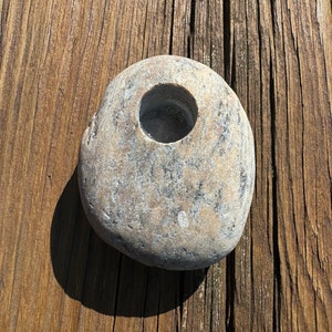 May include: A grey stone with a round hole in the centre. The stone is sitting on a weathered wooden surface.