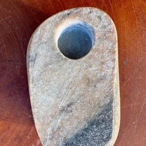 May include: A hand-carved stone incense burner with a large central hole containing a metal mesh screen. The stone is a mix of beige, grey, and black, with a smooth, worn texture. The burner is oval-shaped and sits on a wooden surface.
