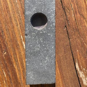 May include: A rectangular, grey stone object featuring a circular mesh screen. The stone has a textured surface and is presented on a wooden surface. The object appears to be a small, decorative item.