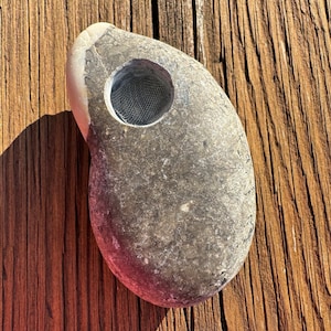 May include: A grey stone pipe with a mesh screen in the bowl. The stone has a smooth, rounded shape with a lighter-coloured tip. The pipe is resting on a weathered wooden surface.