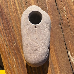 May include: A grey stone with a cylindrical hole. The stone has a natural, organic shape and texture. The hole appears to have a mesh screen inside. The stone is resting on a wooden surface.